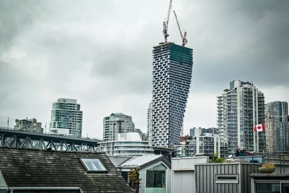 View of Vancouver House skycraper under construction with two cranes at top and other downtown Vancouver buildings surrounding it against a grey sky